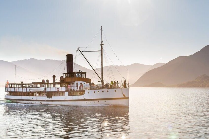 A white paddle steamer with a deck and passengers, sailing on calm waters with mountains in the background. 1