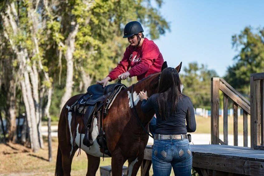 A person riding a horse, another person standing beside the horse, and a wooden fence in the background. 5