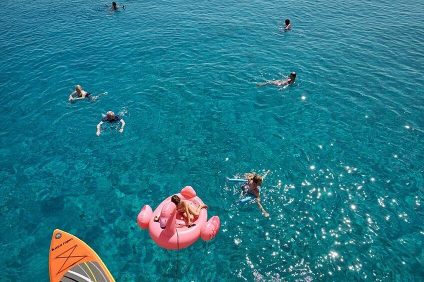 People swimming in clear blue water with a pink inflatable float and a surfboard. 2