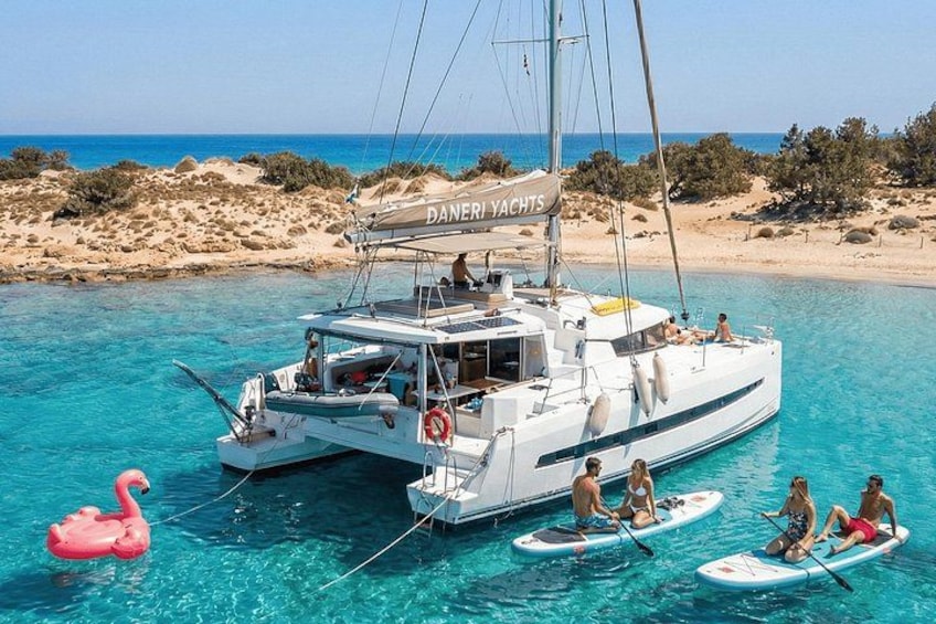 A white catamaran named 'DANERI YACHTS' on clear turquoise water with people on paddleboards and a pink flamingo float. 1