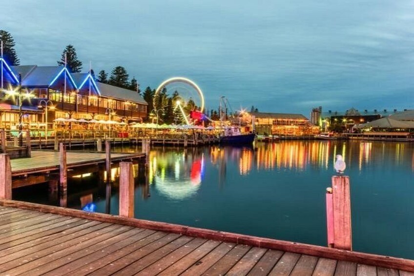 A waterfront with a wooden dock, a large illuminated building, and a circular light structure reflecting on the water. 1