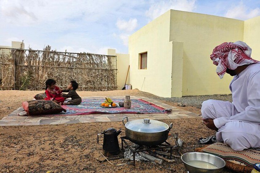 Two people sitting on a mat outdoors, a cooking setup with a pot and a metal container, and a building in the background. 4
