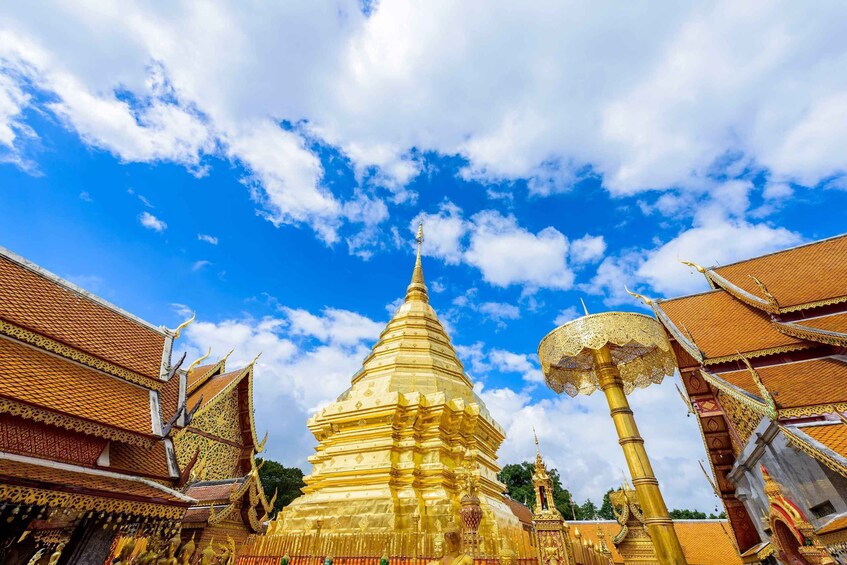 Kultainen stupa, jossa on monimutkaisia koristeita, ja sen ympärillä on perinteistä thaimaalaista arkkitehtuuria kirkkaan sinisen taivaan alla. 2