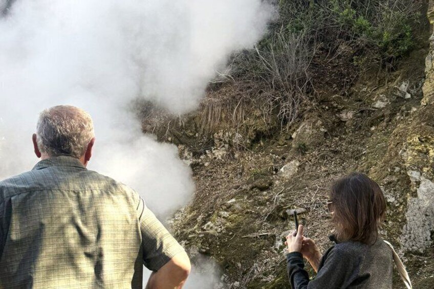 An elderly man and a woman observing steam rising from a rocky hillside. 1