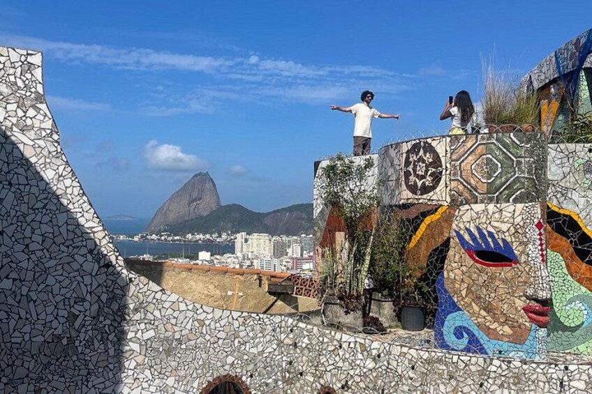 Eine Wand mit einem Mosaik, eine Person darauf stehend, ein farbenfrohes Wandgemälde und eine Aussicht auf Berge und Stadtlandschaft. 2