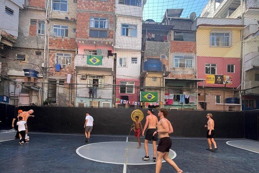 Eine Gruppe von Menschen spielt Basketball auf einem Außenplatz, umgeben von mehrstöckigen Gebäuden mit verschiedenen Schildern und Flaggen. 4