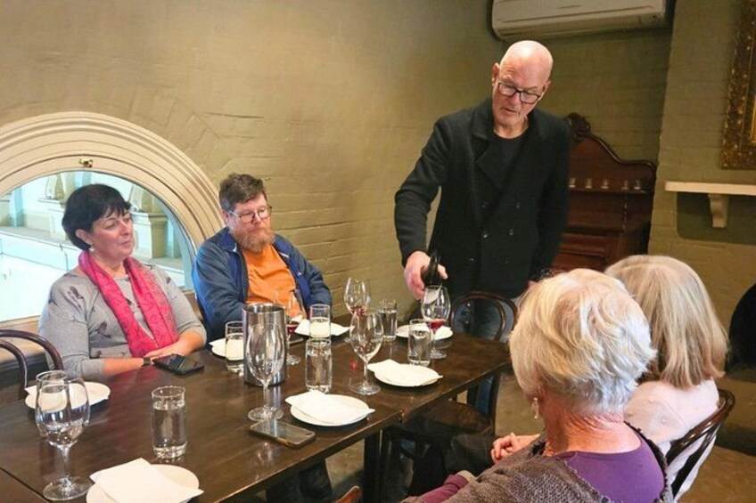 A group of people seated around a table with wine glasses and plates, in a room with a fireplace and a window. 4