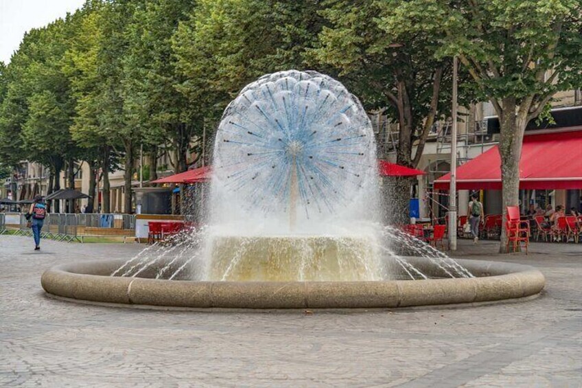 A fountain with a circular water jet in the center, surrounded by a paved area with trees and red umbrellas in the background. 2