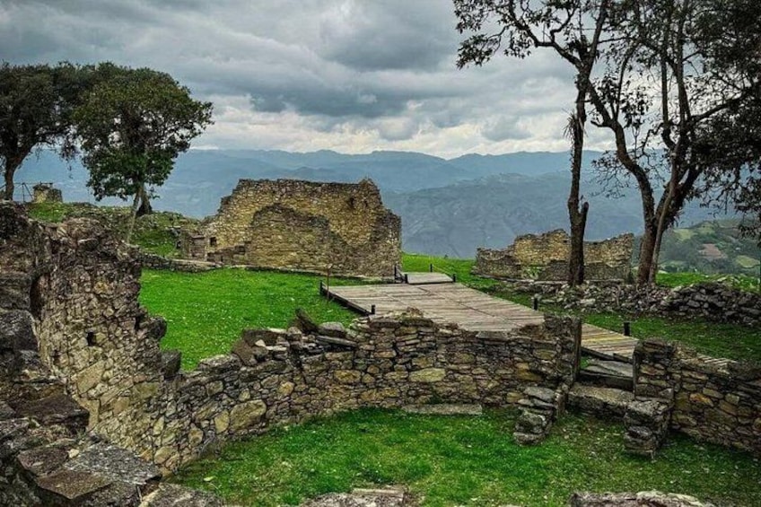 Ancient stone ruins with a grassy pathway, surrounded by trees and mountains in the background. 1