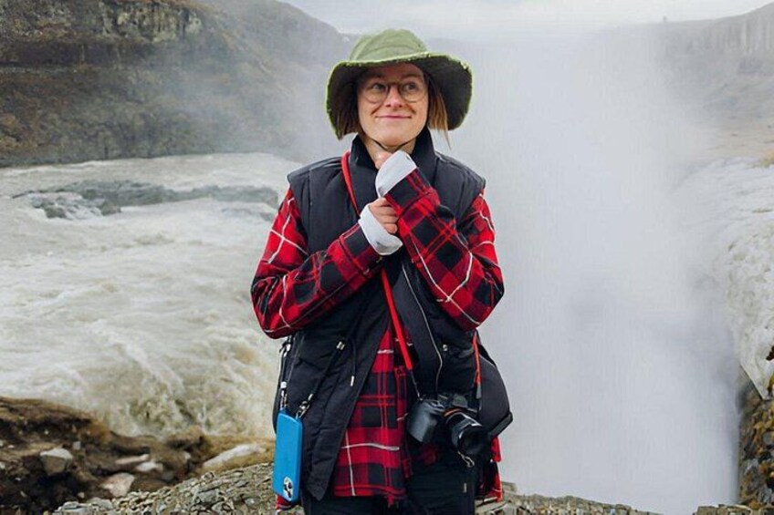 A person standing in front of a waterfall with a camera and a blue bag. 3