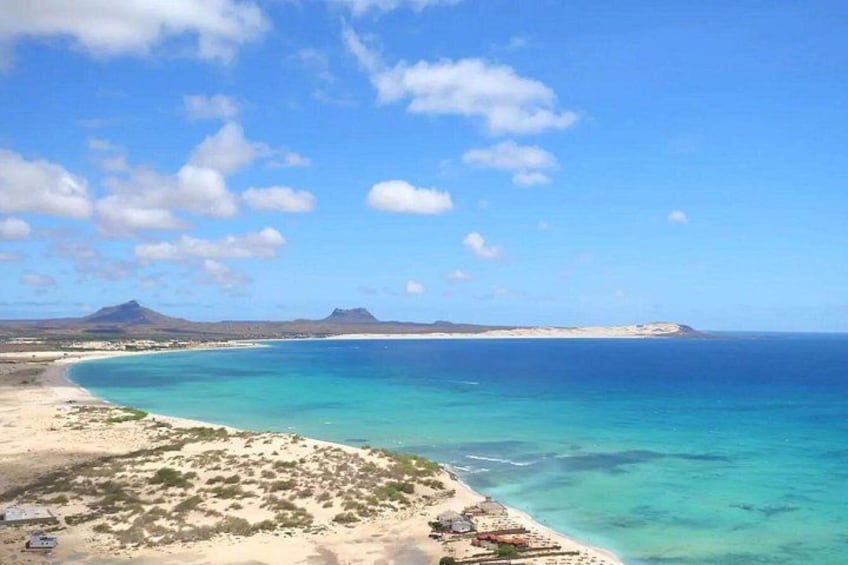 A coastal landscape with a sandy beach, clear turquoise waters, and distant mountains under a partly cloudy sky. 4
