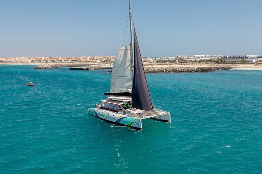A sailboat with a dark sail on turquoise water, a rocky shoreline with buildings in the background. 1