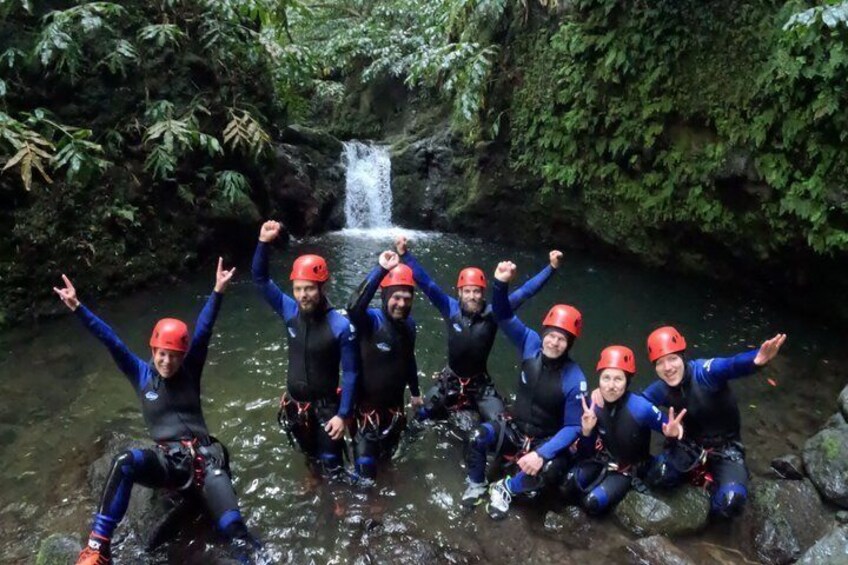 A group of people in wetsuits and helmets posing by a waterfall in a forested area. 3
