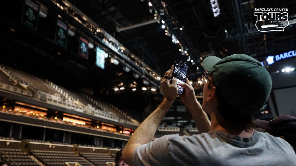 En person tar ett foto i en stor inomhusarena med flera våningar av sittplatser och en väl synlig skylt med texten Barclays Center. 3