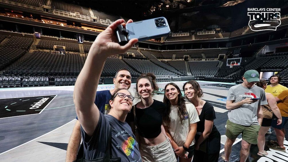 En grupp människor som tar en selfie i en inomhusarena med tomma sittplatser och en basketplan. 1