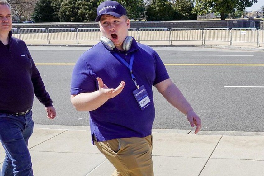A man in a blue shirt and khaki pants walking on a sidewalk with headphones around his neck. 5