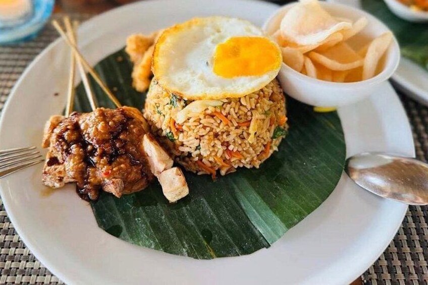 A plate of fried rice with a fried egg on top, accompanied by a side of sambal and a piece of grilled chicken. 1