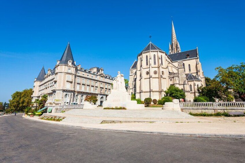 A large historic building with a statue in front, a tall spire, and a paved area in the foreground. 2