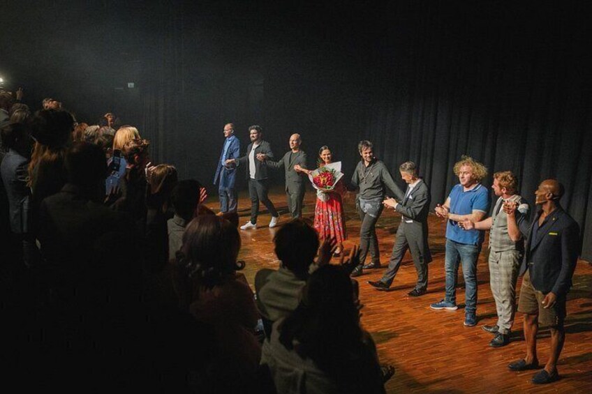 A group of people on a stage with a curtain backdrop, some holding flowers. 3