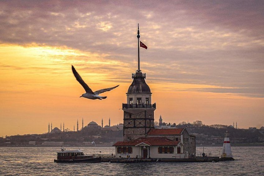 A historic building with a flag, a boat, and a lighthouse by the water during sunset. 3