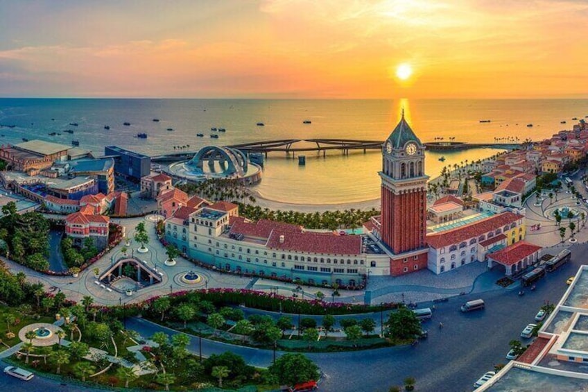 Une vue urbaine d’une ville côtière, avec une tour de l’horloge imposante, un pont et une promenade en bord de mer, le tout baigné par les couleurs du coucher de soleil. 1