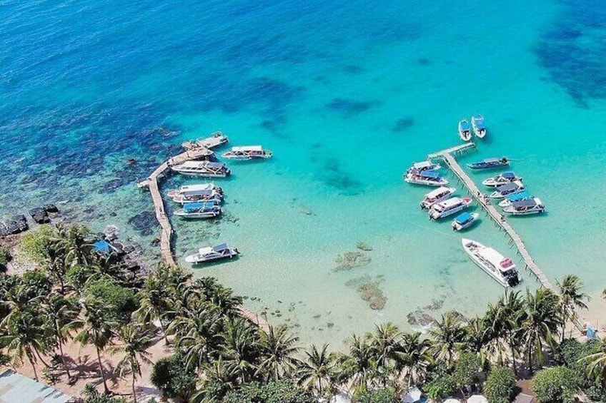 An aerial view of a beach with boats docked at piers, surrounded by palm trees and clear turquoise water. 1