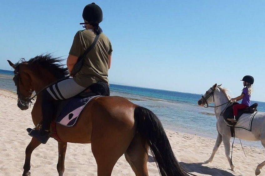 Two people riding horses on a sandy beach with the ocean in the background. 2