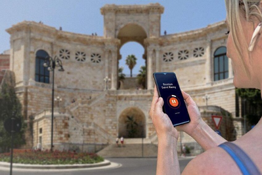 Una persona usando un teléfono inteligente con un icono de aplicación etiquetado 'Baston Saint Damien' frente a un edificio histórico de piedra con arcos y detalles decorativos. 5