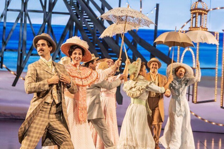 A group of people in vintage costumes holding umbrellas on a stage with a backdrop of a bridge and water. 1