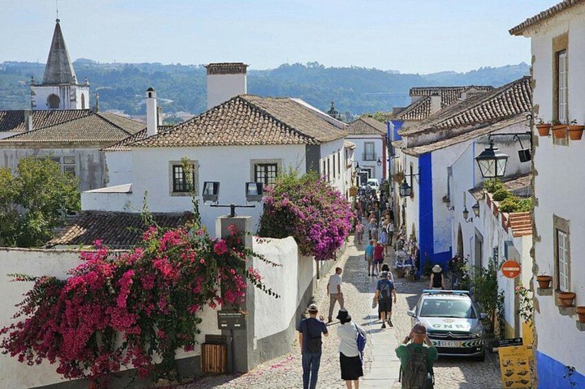 A narrow street with white buildings, a church spire, and people walking. 2