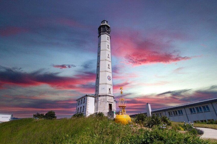 A lighthouse with a white tower and a red top, surrounded by greenery and a building with a white facade. 4