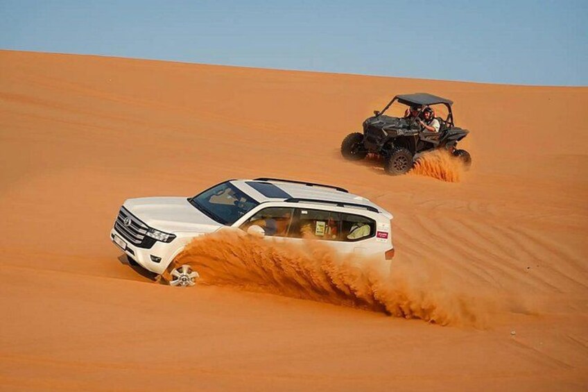 A white SUV and a black ATV driving through a desert with sand dunes. 1