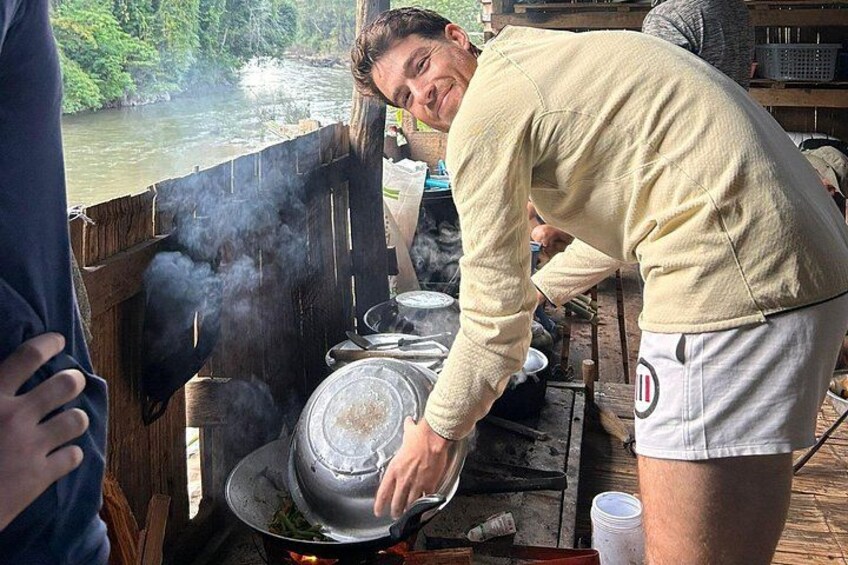 A person cooking outdoors with a large pot on a stove, surrounded by wooden structures and a river in the background. 5