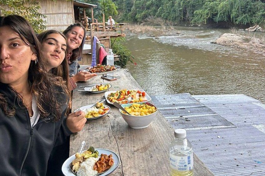 Three people dining at a wooden table by a river. 2