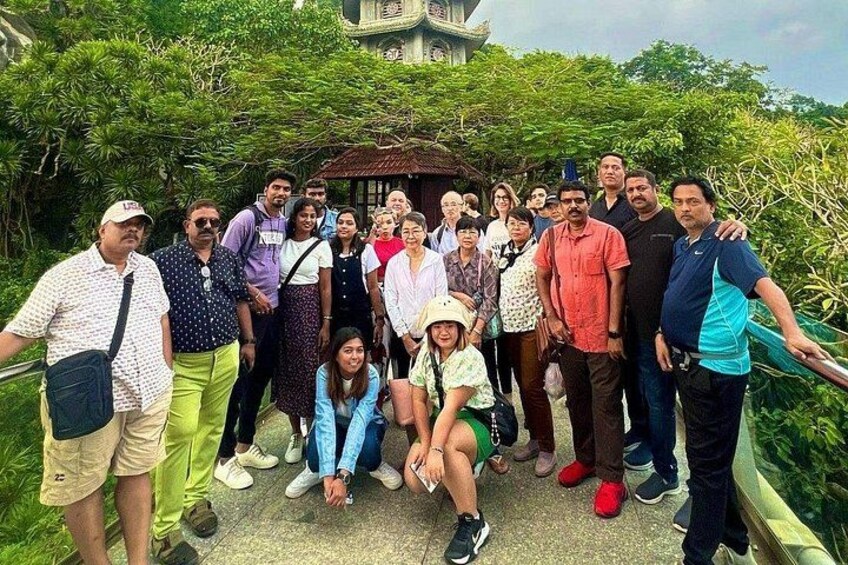 A group of people posing in front of a traditional building with a tiled roof, surrounded by greenery. 1