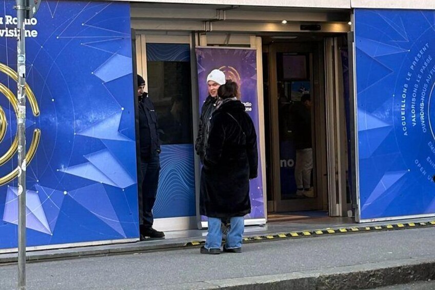 Ein Gebäudeeingang mit einem blauen, geometrischen Muster an der Wand, einer Glastür und einer Person davor. 1