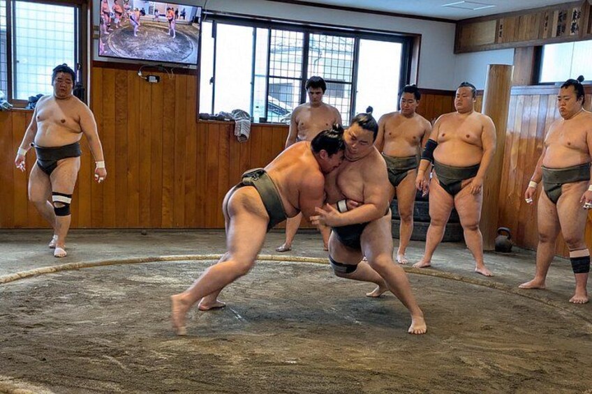 A sumo wrestling match with wrestlers in traditional attire, a wooden floor, and spectators in the background. 1