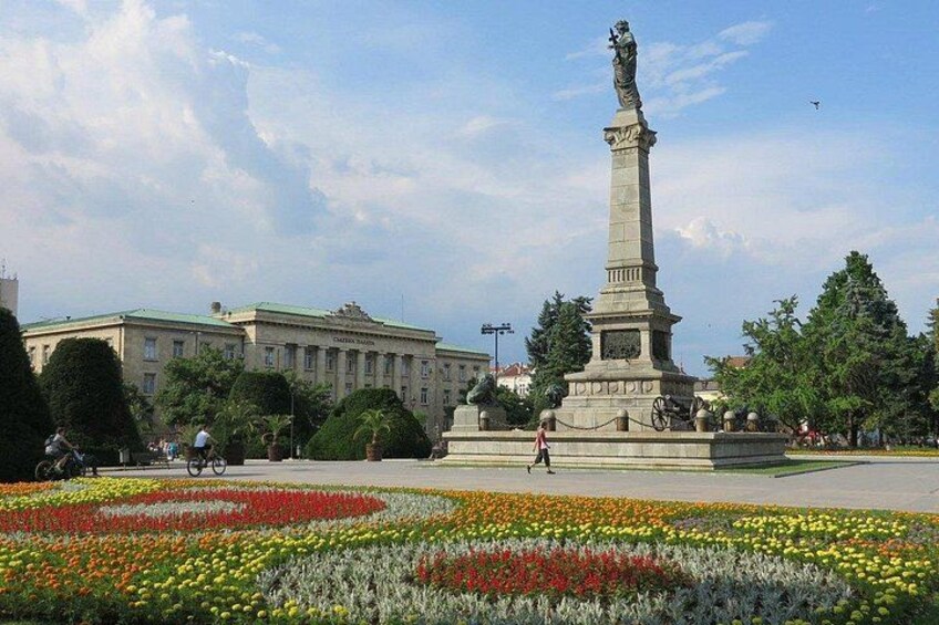 A monument in a park with flower beds and a building in the background. 2