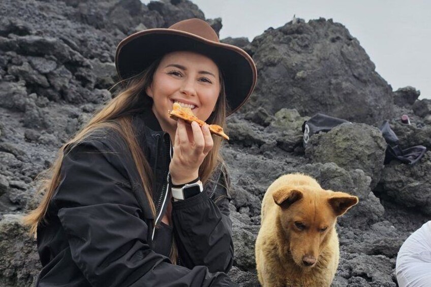 A person wearing a black jacket and brown hat eating pizza, with a dog nearby, on a rocky terrain. 1