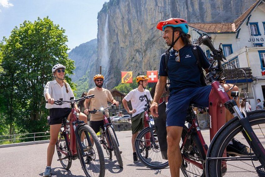 A group of cyclists with mountain bikes, a mountain backdrop, and a building with a sign. 3