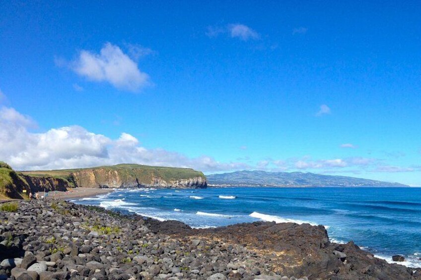 A rocky coastline with waves crashing onto the shore, a clear blue sky, and distant hills. 1