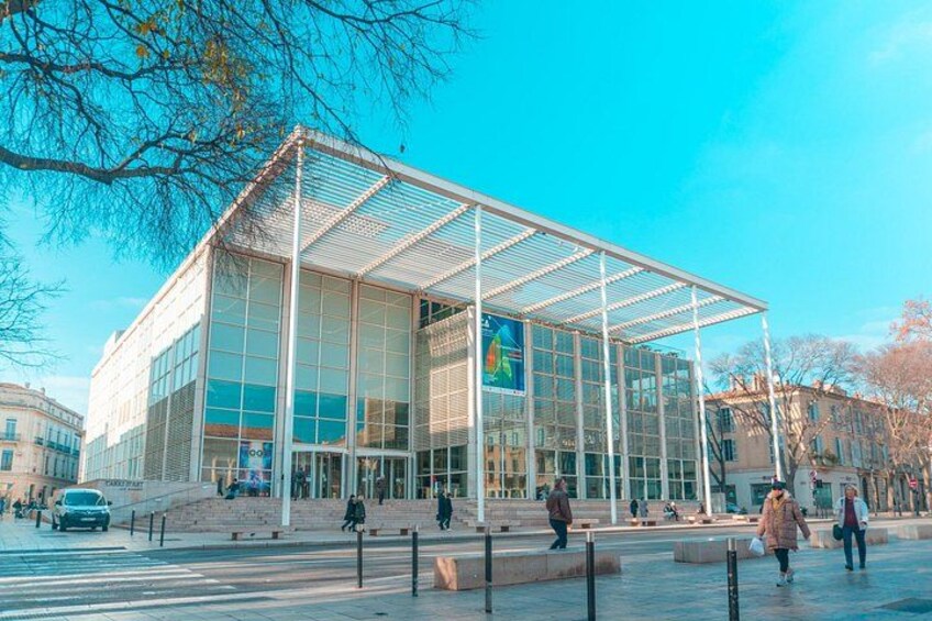 A modern glass building with a metal framework, surrounded by a plaza with benches and a few pedestrians. 4