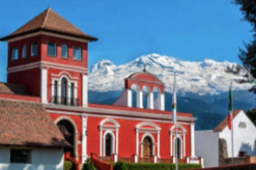 A red church with a bell tower, a snow-capped mountain in the background, and a flagpole with flags. 1