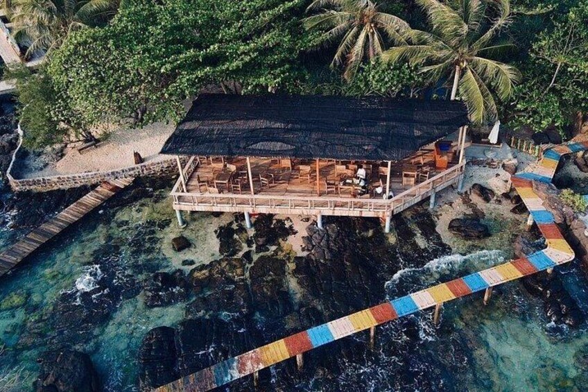 A wooden structure with a thatched roof on stilts over water, surrounded by palm trees and a colorful bridge. 3