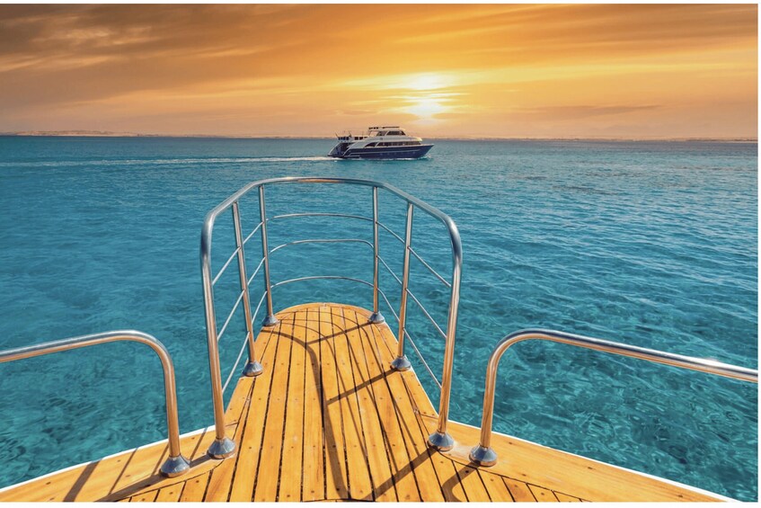 A wooden boat deck with metal railings, clear turquoise water, and a yacht in the distance during sunset. 1
