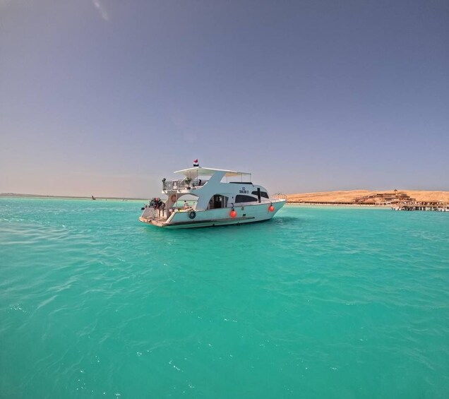 A white boat with a flag on top, floating on turquoise water near a sandy beach with buildings in the background. 4