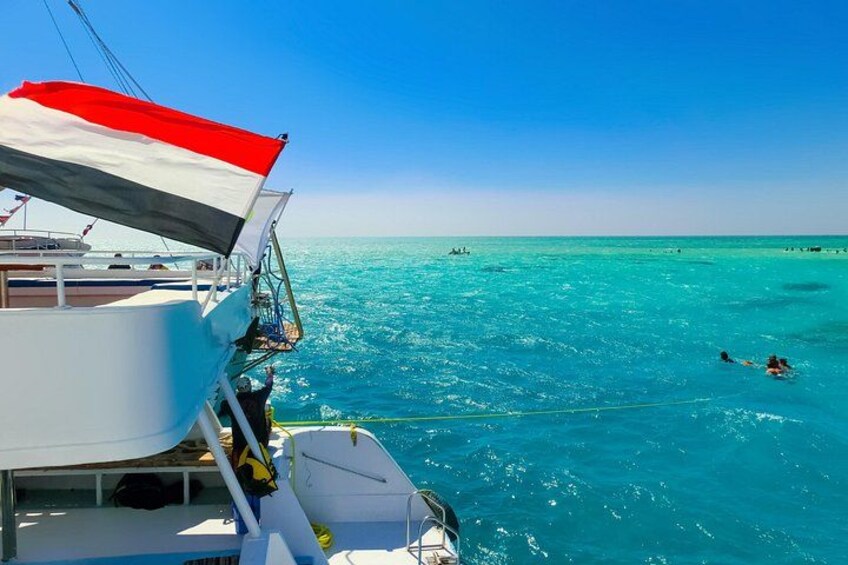 A boat with a flag, clear turquoise water, and people swimming. 2
