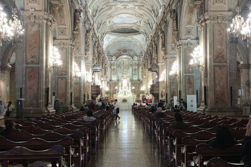 Ein reich verzierter Kirchenraum mit Reihen von Kirchenbänken, einer hohen Decke mit aufwendigen Verzierungen und Kristalllüstern. 4