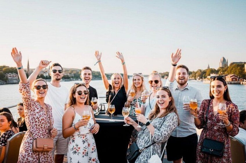 A group of people on a boat, holding drinks, with a body of water and buildings in the background. 1