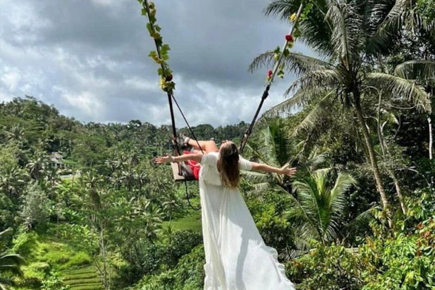 Una persona con un abito bianco dondola su un'altalena circondata da vegetazione rigogliosa e palme. 1